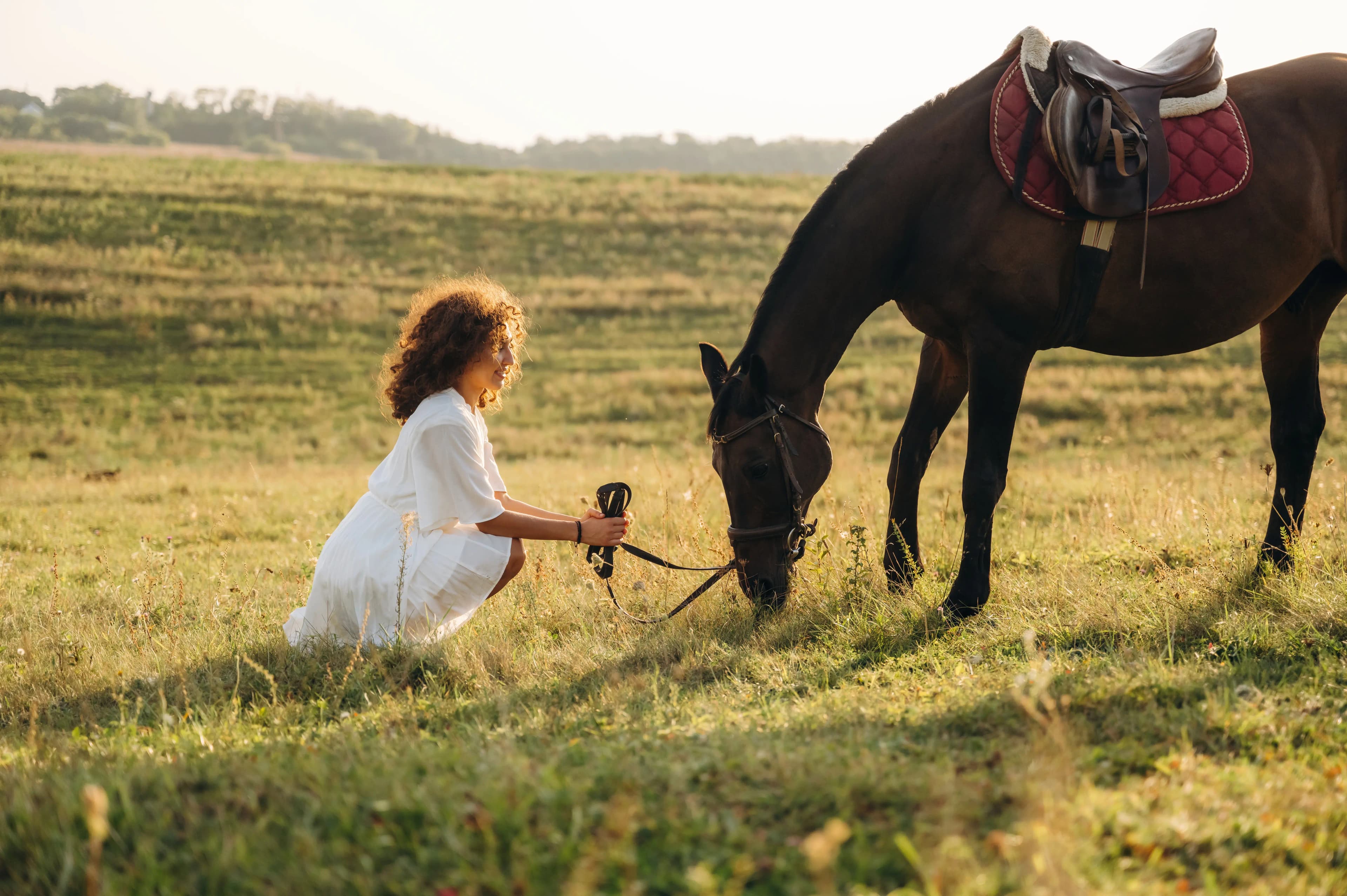 A woman sitting quietly beside her horse as it grazes in a meadow