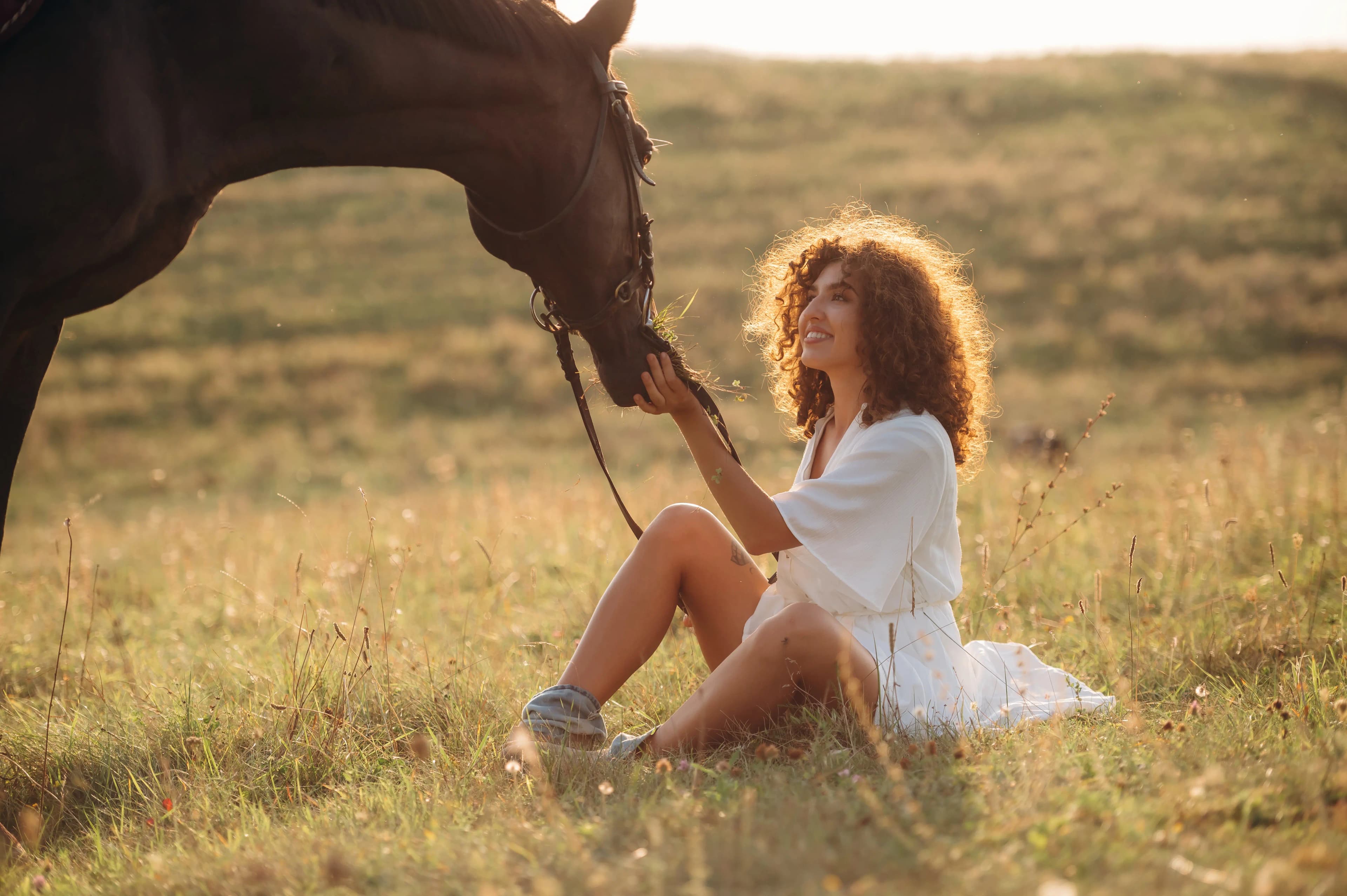 A woman sitting peacefully beside her horse in a sunlit field